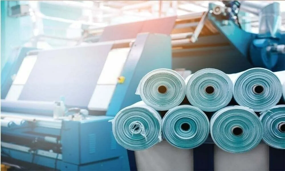 Rolls of finished fabric beside a knitting machine on the mill floor